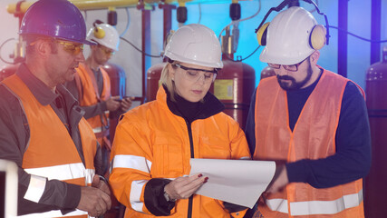 Lady inspector with engineers in colorful uniform and white helmets reads paper documents at contemporary industrial complex close view