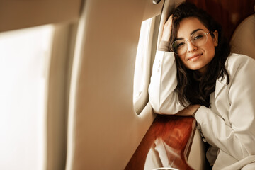 Young, beautiful brunette girl with big glasses walks in the passenger seat in a charter plane or business jet near the porthole and smiles