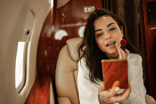 Young Brunette In A White Suit Corrects Makeup With A Mirror In Powder Flying Aboard A Business Jet Plane