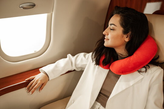 Rest in the plane. A young girl in a white suit is resting and looking in the window of a business plane, using a red neck pillow for travel