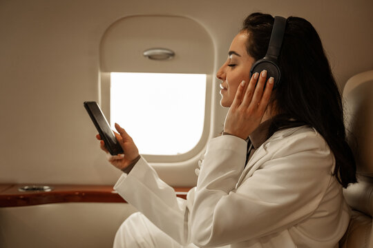 Brunette girl in a white suit, a passenger of the plane, relaxes in flight, listening to music and watching videos on the phone, listening to the sound through wireless headphones