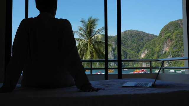 Man in bad mood, cant start or continue work, sit still and stare out to tropical bay. Silhouetted shot of tired freelancer against window. Open laptop computer next to guy on the bed