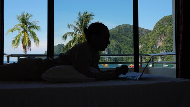 Man lie on bed, work using laptop, silhouetted shot against window. Green tropical nature and sea bay seen on background. Stereotyped image of freelancer lifestyle