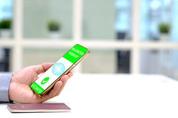 A man holding a smart phone with a digital illustration of a certificate of vaccination against the Covid-19 disease and passport on white table.