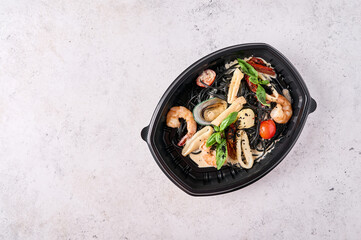 Black spaghetti Nero with seafood, tomatoes and basil in black plastic container on light background. Designer food concept for take away or delivery