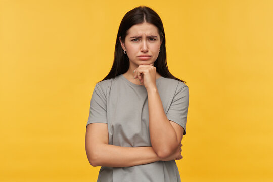 Pensive Concentrated Brunette Young Woman In Gray Tshirt Keeps Hands Folded And Thinking Over Yellow Background Looking At Camera