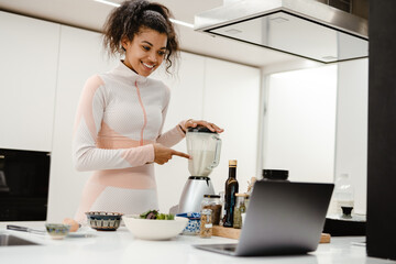 Black woman pointing finger at blender and using laptop while cooking