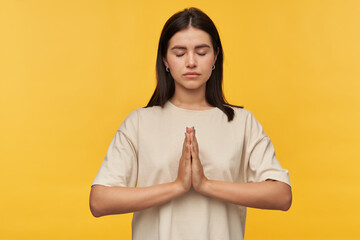 Obraz premium Peaceful beautiful young woman with dark hair and eyes closed in white tshirt keeps hands in praying position and meditating over yellow background