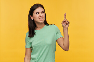 Angry displeased brunette young woman in mint tshirt looks irritated and pointing up by finger over...