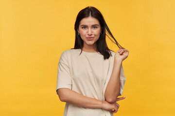 Obraz premium Studio portrait of smiling attractive young woman in white tshirt standing and touching her dark hair over yellow background Looking at camera