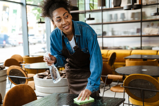 Black waitress wearing apron cleaning table while working in cafe - Powered by Adobe