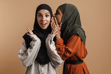 Young muslim woman in hijab whispering secret to her surprised friend