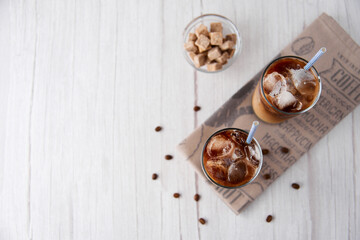 Iced coffee latte cappuccino in a tall glass with cream or milk and beans, straws on light background. Cold tasty summer refreshment beverage concept. Selective focus, copyspace. Flat lay, top view.