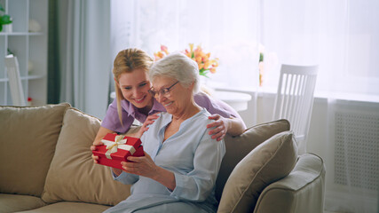 Long haired blonde in purple dress gives small box with present to aged lady
