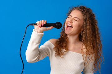 Young joyful curly-haired woman singing in microphone against blue background