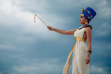Woman in image of egyptian queen Nefertiti stands against sky with whip.