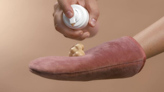 Dosing bronzing lotion or tanning cream from a flask with a doser to a pink tan applicator glove. High quality close-up slow motion studio video footage beige background.