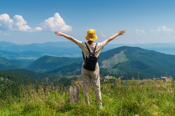 A girl traveler with a backpack stands with her arms outstretched in the mountains in summer. The top of the mountain evokes good or uplifting mood