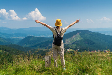 Fototapeta premium A girl traveler with a backpack stands with her arms outstretched in the mountains in summer. The top of the mountain evokes good or uplifting mood