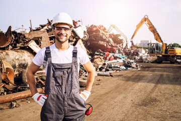 Portrait of worker standing in metal junk yard with crane lifting scrap metal for recycling. - obrazy, fototapety, plakaty