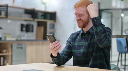 Redhead Man Celebrating Success on Smartphone in Cafe 