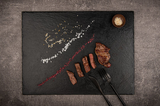 Grilled meat is placed on a black stone dish. Next to the knife, salt and pepper shaker.
