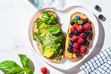 Peanut butter and berries toast and avocado toast with basil on heart plate. Healthy vegan breakfast concept.