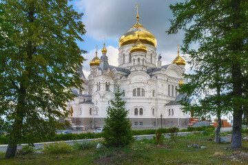 Fototapeta na wymiar View of the ancient church in the Belogorsk monastery (Perm region, Russia) on a cloudy summer day. White stone temple with golden domes against the backdrop of a cloudy sky and tall trees 