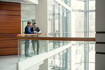 Two businessmen discussing document together while standing in the corridor of the modern office building