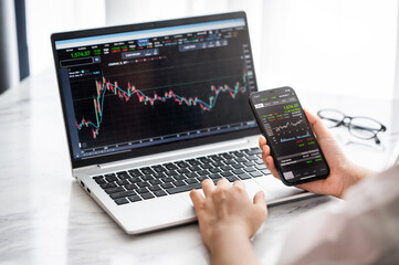Hand holding smartphone with stock market data and using laptop display graph and chart for analyze and check before trading stocks online