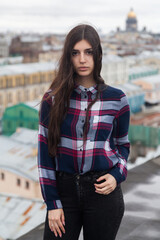 An Armenian girl with long black hair in a plaid shirt stands on a rooftop in the center of St. Petersburg