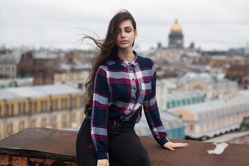 An Armenian girl with flowing long black hair in a checkered shirt sits on a rooftop in the center of St. Petersburg