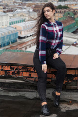 An Armenian girl with flowing long black hair in a checkered shirt sits on a rooftop in the center of St. Petersburg