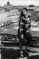 An Armenian girl with flowing long black hair in a checkered shirt sits on a rooftop in the center of St. Petersburg