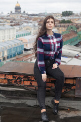 An Armenian girl with flowing long black hair in a checkered shirt sits on a rooftop in the center of St. Petersburg