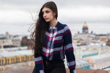 portrait of an Armenian girl with fluttering long black hair in a checkered shirt and jeans on a rooftop in the center of St. Petersburg