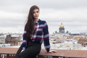 Fototapeta premium An Armenian girl with flowing long black hair in a checkered shirt sits on a rooftop in the center of St. Petersburg