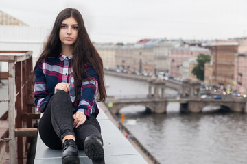 An Armenian girl with fluttering long black hair in a checkered shirt and jeans sits on the edge of...