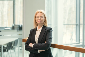 Portrait of confident mature businesswoman in suit standing with her arms crossed in the office corridor and looking at camera