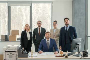 Portrait of group of business people looking at camera while standing at office with their leader in the centre at the table