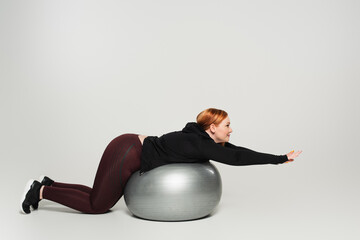 Side view of plus size woman working out on fitness ball on grey background