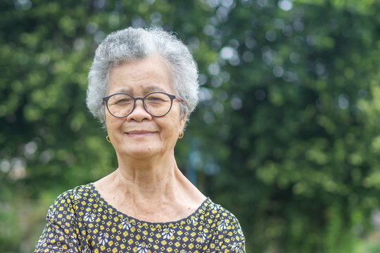 Portrait of an elderly Asian woman with short gray hair, wearing glasses, smiling and looking at the camera while standing in a garden. Aged people and relaxation concept