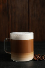 Glass cup of delicious layered coffee and beans on black table against wooden background, space for text