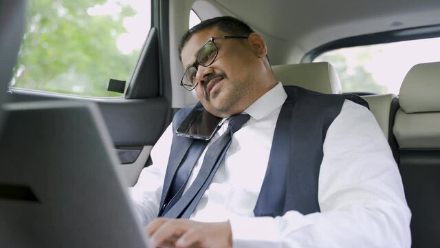 Young Indian business man busy tlking on phone and working on laptop while travelling in car - concept of successful business people, busy schedule and meetings.