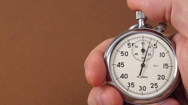 Human Hand hold classic steel mechanical analog stopwatch on brown background. Hand holding a stopwatch against a brown background. Man trainer pushing a stopwatch button.