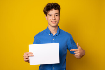 Beautiful european schoolboy in blue shirt posing with blank white paper against yellow background.