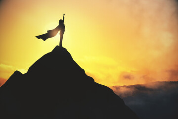 Success and satisfaction concept with super woman in waving coat on top of dark rock on yellow foggy sky background. - obrazy, fototapety, plakaty
