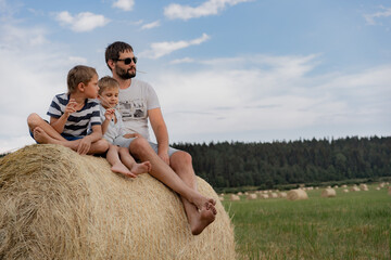 portrait of man and his two little sons sitting on round haystack in a green field on sunny summer day.