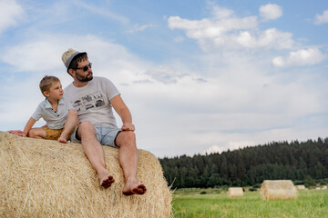 portrait of man and his little son sitting on round haystack in a green field on sunny summer day.