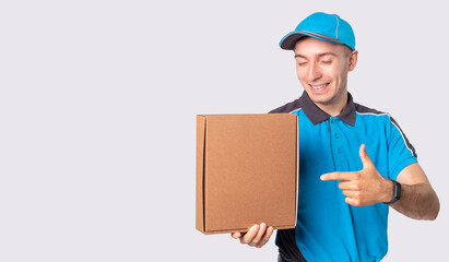 Smiling courier in blue uniform and a cap is pointing to the parcel he must deliver it to destination.